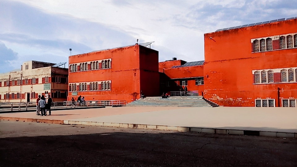 Rust-red school building with wide forecourt and a small group of arriving students
